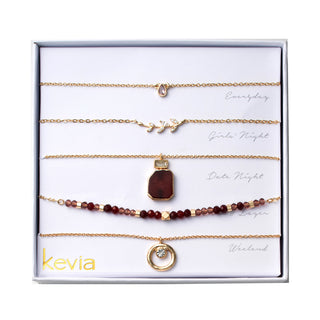 Five piece necklace set packaged in a gift box featuring a gold bezel set crystal solitaire necklace, a gold curved crystal leaf motif necklace, a rectangular deep red stone pendant necklace with a small rectangular crystal accent, a faceted garnet bead necklace with gold spacer beads, and a gold open circle pendant necklace with a centered clear crystal.