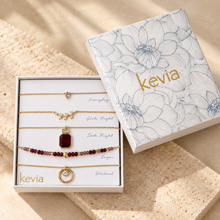 Five-piece gold necklace set with red stones, beads, and crystal pendants in an open box beside a floral Kevia lid on cream stone.