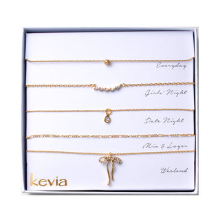 Five-piece gold necklace set featuring a fine chain with a small crystal accent, a crystal cluster station necklace, a bezel-set crystal solitaire pendant necklace, a mixed-link chain necklace, and a pavé crystal bow pendant necklace, packaged in a gift box.
