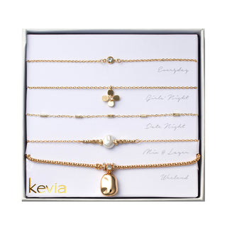 Five-piece gold necklace set featuring a bezel-set crystal solitaire necklace, polished clover charm necklace, station chain with slim rectangular accents, pearl bead necklace with gold spacers, and a chunky chain necklace with an organic pebble pendant and crystal spacer, packaged in a gift box.
