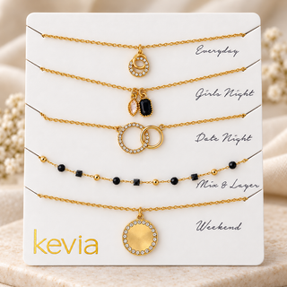 Five gold-toned layered necklace sets displayed on a white card labeled kevia, with handwritten style labels: Everyday, Girls Night, Date Night, Min & Layer, and Weekend. The necklaces feature different pendant and bead designs, including clear rhinestone accents and black beads.