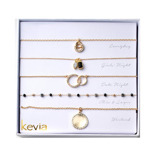 Five-piece gold necklace set featuring a pavé open circle pendant necklace, paired charm pendant necklace with a light stone and black stone charm, interlocking pavé circle pendant necklace, station chain necklace with mixed dark and neutral bead accents, and a round pavé disc pendant necklace, packaged in a gift box.