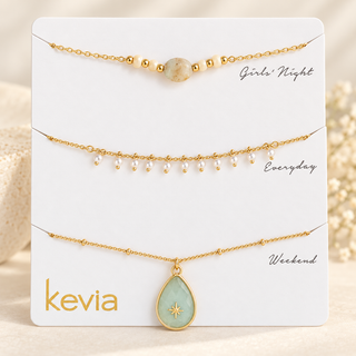 Three gold-toned necklace sets displayed on a white card labeled kevia, with handwritten style labels Girls' Night, Everyday, and Weekend. The top set has a round stone pendant, the middle has small dangling beads, and the bottom has a larger teardrop pendant with a pale green stone and gold star detail.