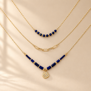Three layered gold necklaces with deep blue beads, displayed on a warm beige background with soft shadow overlays. The top necklace has a short curved section of blue beads, the middle has a paperclip link chain, and the bottom features a V-shaped arrangement with blue beads, gold spacers, and a small clear stone heart pendant.