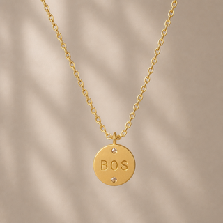 Gold chain necklace with a round pendant stamped BOS and a small crystal accent.