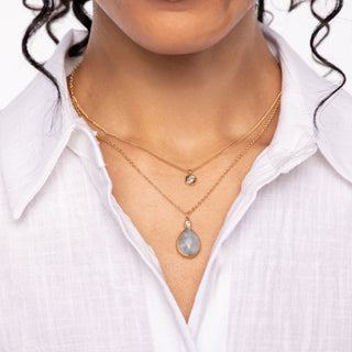 Model wearing two gold layered necklaces, one with a small bezel-set round charm and the other with a faceted teardrop pendant.