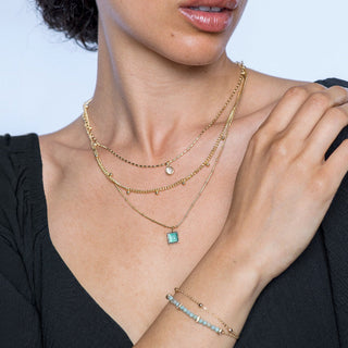 Woman wearing layered gold necklaces and a bracelet with a blue gemstone on a light blue background.
