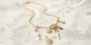 Gold paperclip chain necklace with holiday charms including snowflake, bow, star, and emerald stone on soft winter fabric.