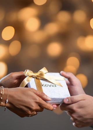 Two people exchanging a wrapped jewelry gift box featuring a curated bundle, shown in warm holiday lighting for the Kevia gift collection hero image.