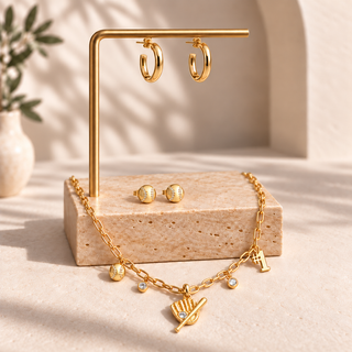 Gold jewelry set displayed on a stone stand, including hoop earrings hanging from a vertical bar, stud earrings on the base, and a chain necklace with small charms and a fan-shaped pendant.
