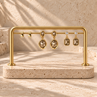 A gold jewelry display stand holding three pairs of earrings, including oval and rectangular smoky gemstone drops, set against a warm beige stone background with soft shadows.

