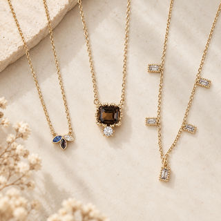 Three gold necklaces with unique pendant designs are displayed on a textured beige surface with dried flowers. One necklace features a small blue and white leaf pendant, the center necklace has a square brown gemstone pendant with a clear round accent, and the right necklace has a Y-shaped design with rectangular clear gemstones.