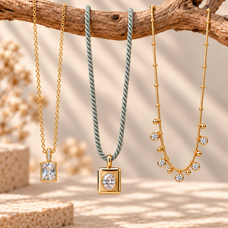 Three gold-tone necklaces displayed on a rustic wooden branch: one with a rectangular clear gemstone pendant, one with a round clear gemstone pendant on a twisted sage green cord, and one with a beaded gold chain and small clear gemstone accents.